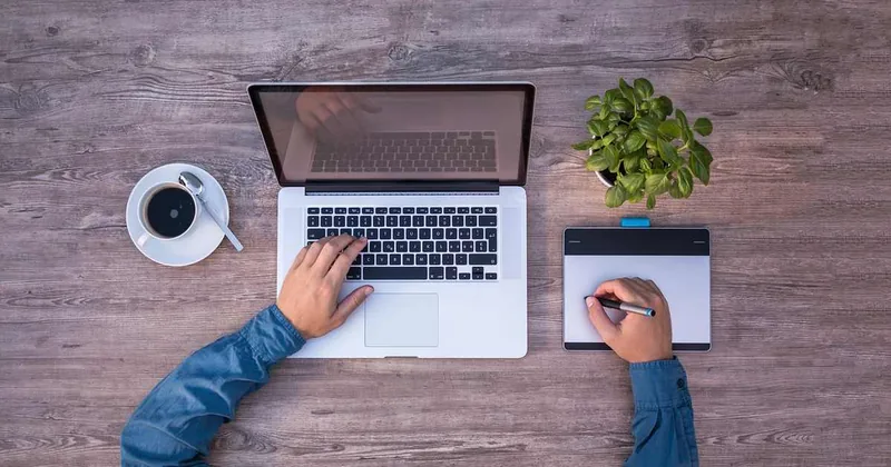 Des mains sur un ordinateur portable et une tablette à écrire, avec une tasse de café et une plante verte sur une table en bois