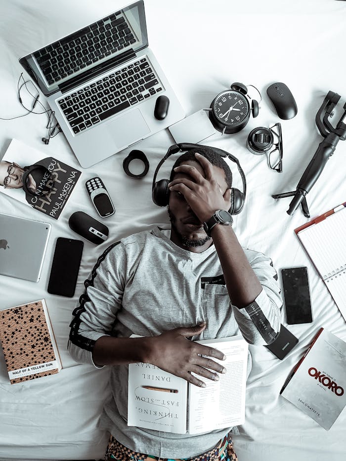 gallery-02 A young man lying on a bed surrounded by modern gadgets, books, and tech devices, illustrating digital lifestyle.